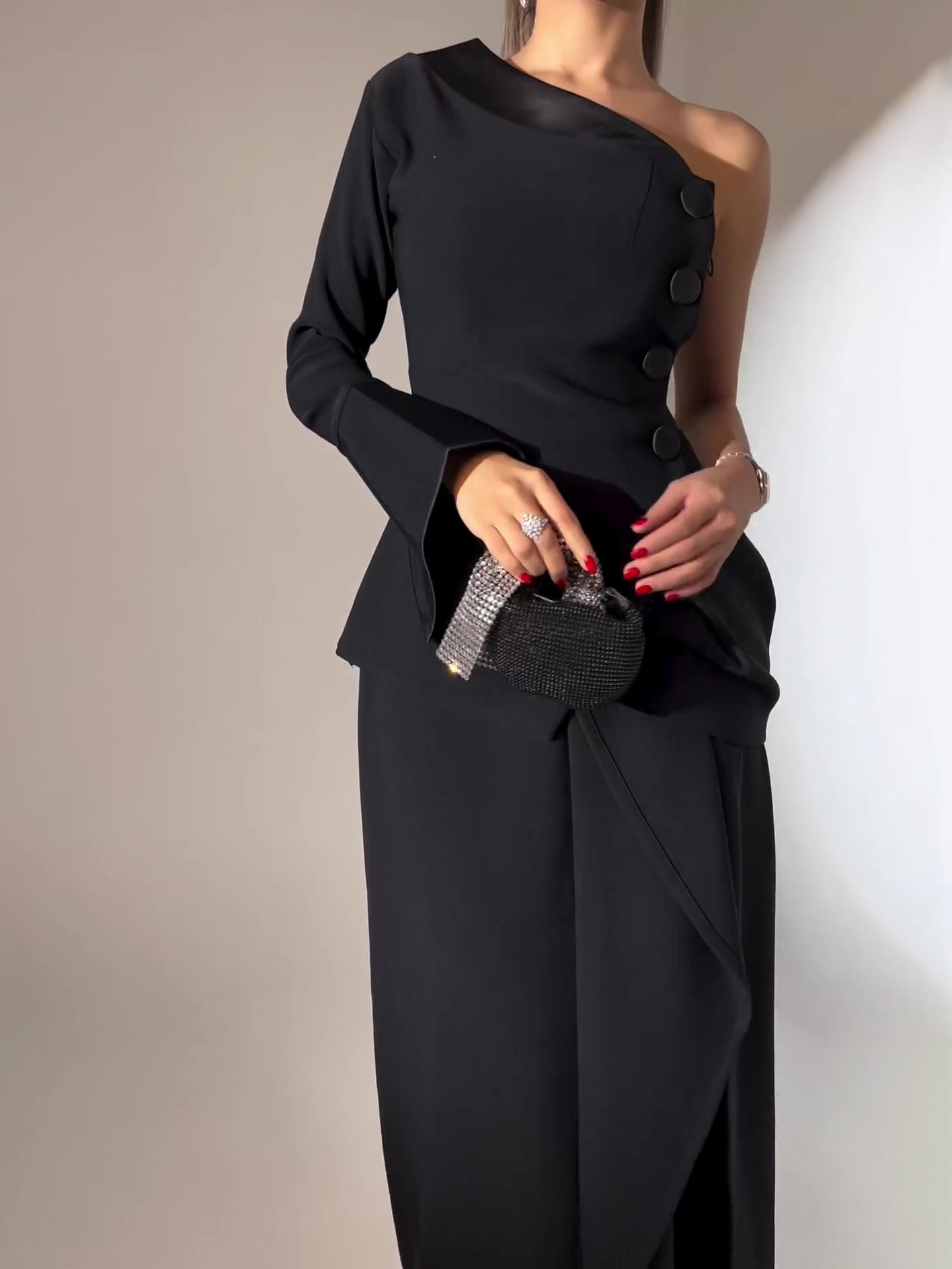 Black one-shoulder dress worn by a person holding a small black clutch against a neutral background