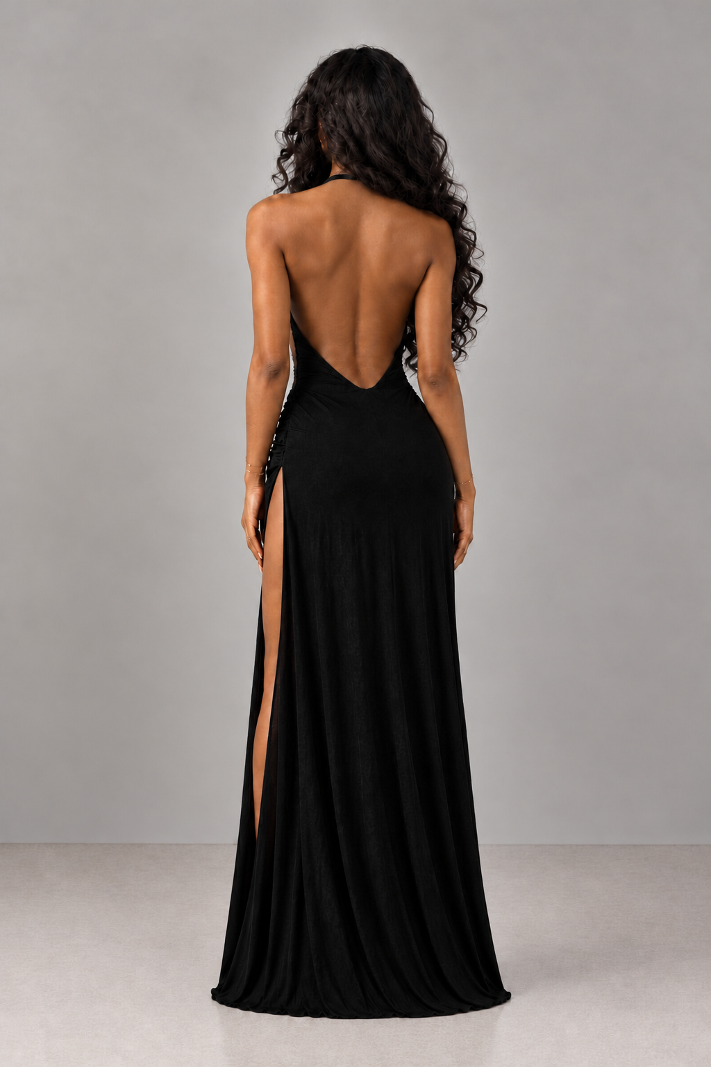 A woman shown from the back wearing a floor-length black gown with a deep open back and a high side slit, photographed against a plain light gray studio background.