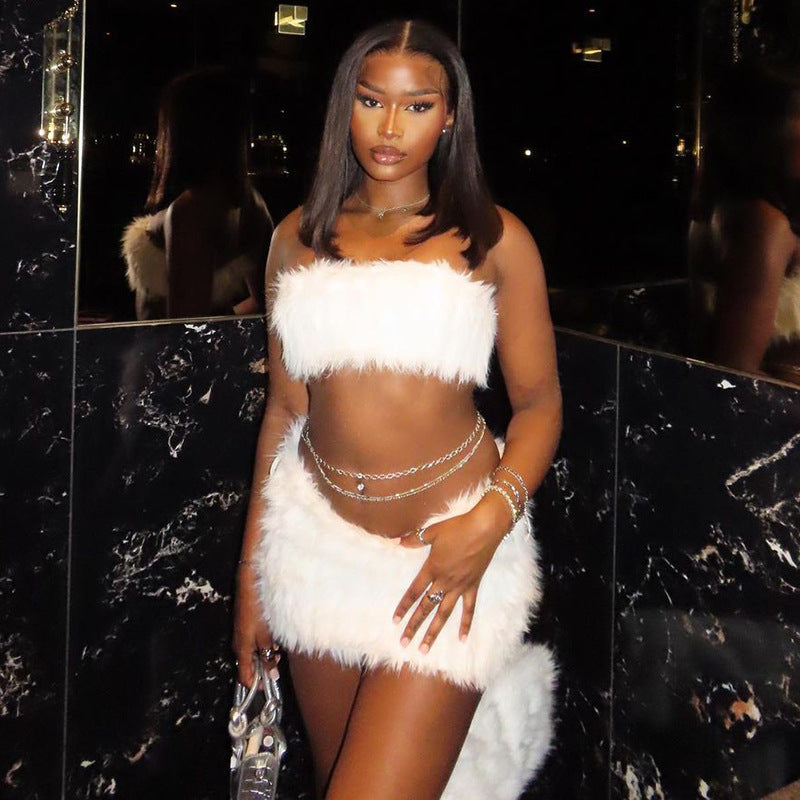 Woman in a white feathered outfit tube top and mini skirt standing against a black marble wall.