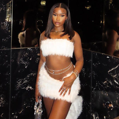 Woman in a white feathered outfit tube top and mini skirt standing against a black marble wall.