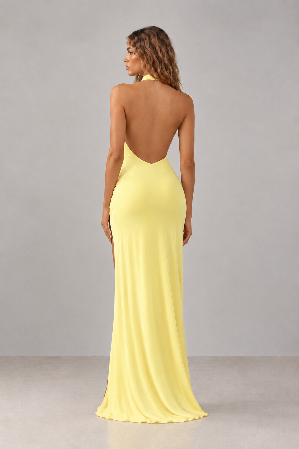 A woman shown from the back wearing a floor-length yellow gown with an open back and side slit, photographed in a studio against a light gray background.