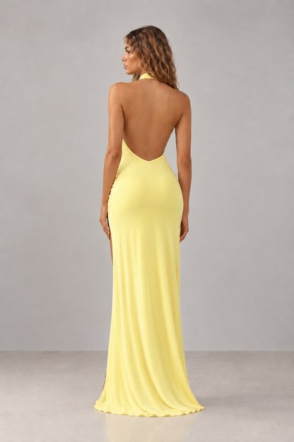 A woman shown from the back wearing a floor-length yellow gown with an open back and side slit, photographed in a studio against a light gray background.