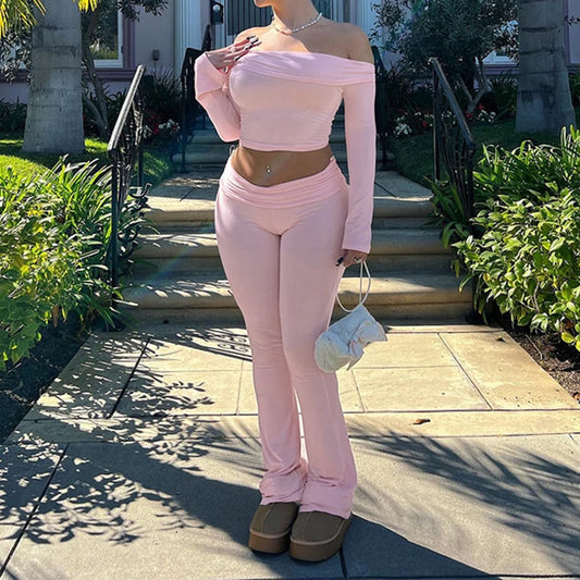 A person wearing an off-shoulder long sleeve top and flare pants in pink, standing on a sidewalk with plants in the background.