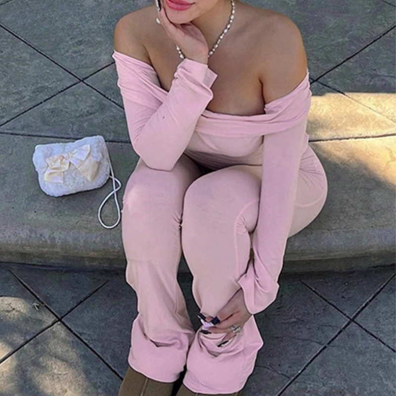 A person wearing an off-shoulder long sleeve top and flare pants in pink, standing on a sidewalk with plants in the background.