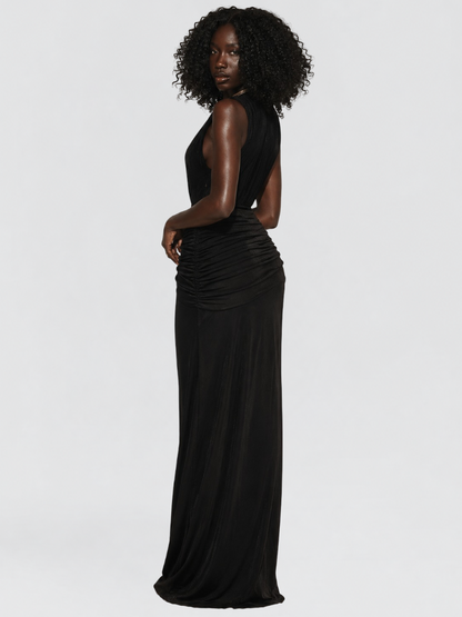 A woman modeling a sleeveless black maxi dress with a draped ruched waist, shown from a side-back angle against a light gray studio background.