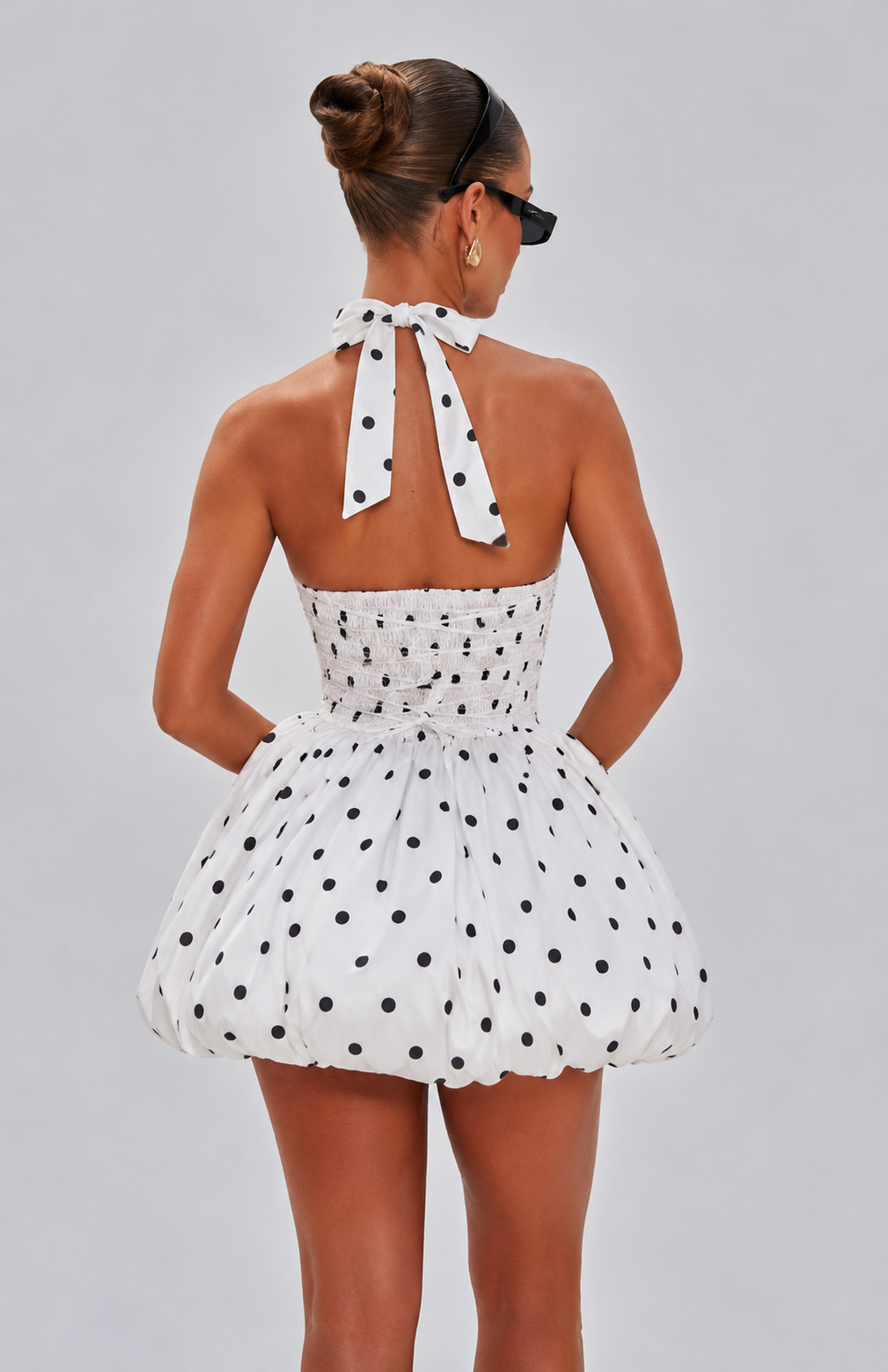 A woman shown from the back wearing a white halter mini dress with black polka dots, featuring a smocked bodice and a voluminous ruffled skirt, set against a solid gray studio background.