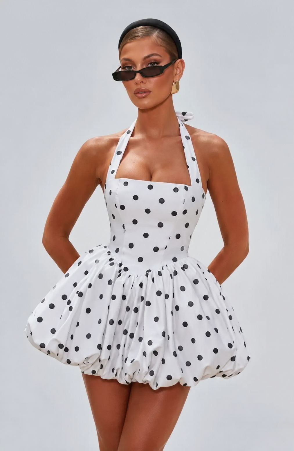 A woman modeling a white halter mini dress with black polka dots, styled with black sunglasses and black high-heeled sandals against a solid gray studio background.