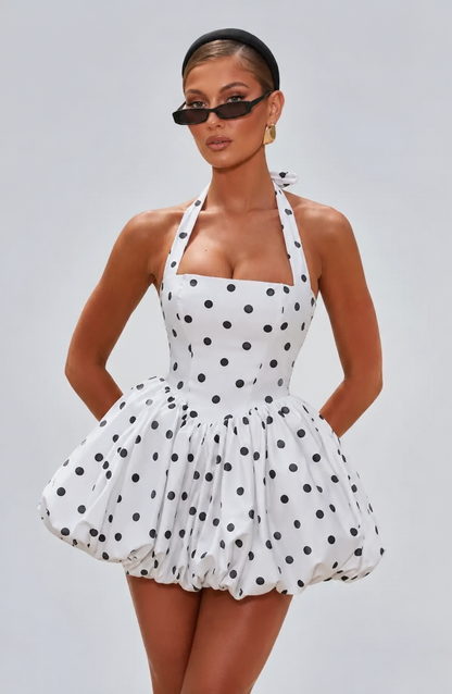 A woman modeling a white halter mini dress with black polka dots, styled with black sunglasses and black high-heeled sandals against a solid gray studio background.
